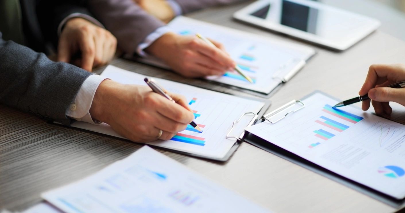 Hands of multiple people pointing at and reviewing charts and graphs on a table, representing teamwork, collaboration, and data analysis in project management.